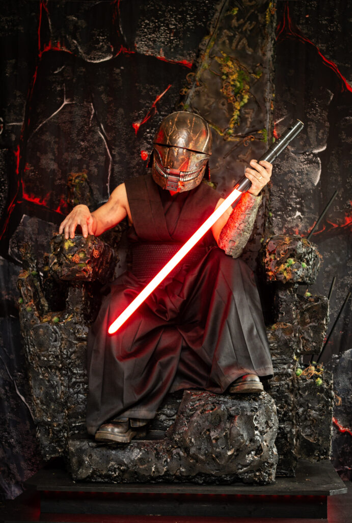 Image of Newry Bakri dressed up in cosplay on a throne holding a red light sabre in his left hand.