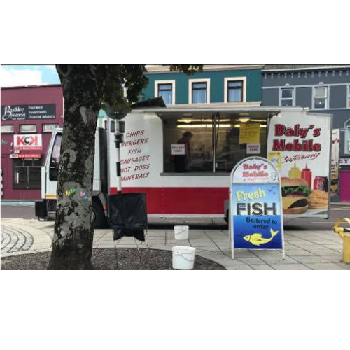 Picture of John Daly's chip van parked up on the side of a street in Dunmanway. There is a sign with Fish outside the front of the chip van. A tree is the the left of the picture. John Daly is working inside the chip van.